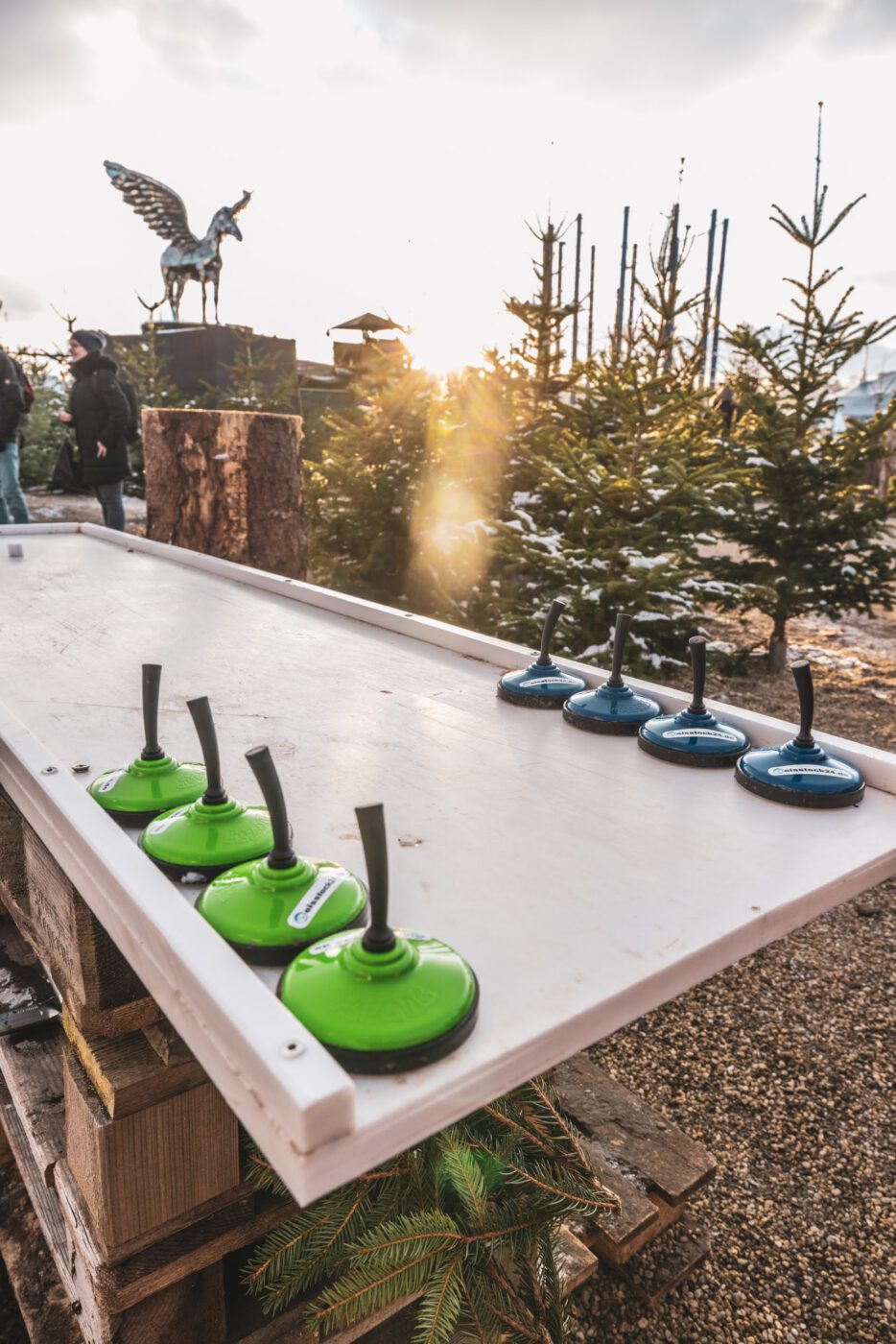 Expertentipps für Eisstockschießen als Weihnachtsfeier - zu sehen ist eine kleine Nachbildung einer Eisstockbahn mit Mini-Stöcken. Im Hintergrund sieht man den Zauberwald auf dem Tollwood Winterfestival.