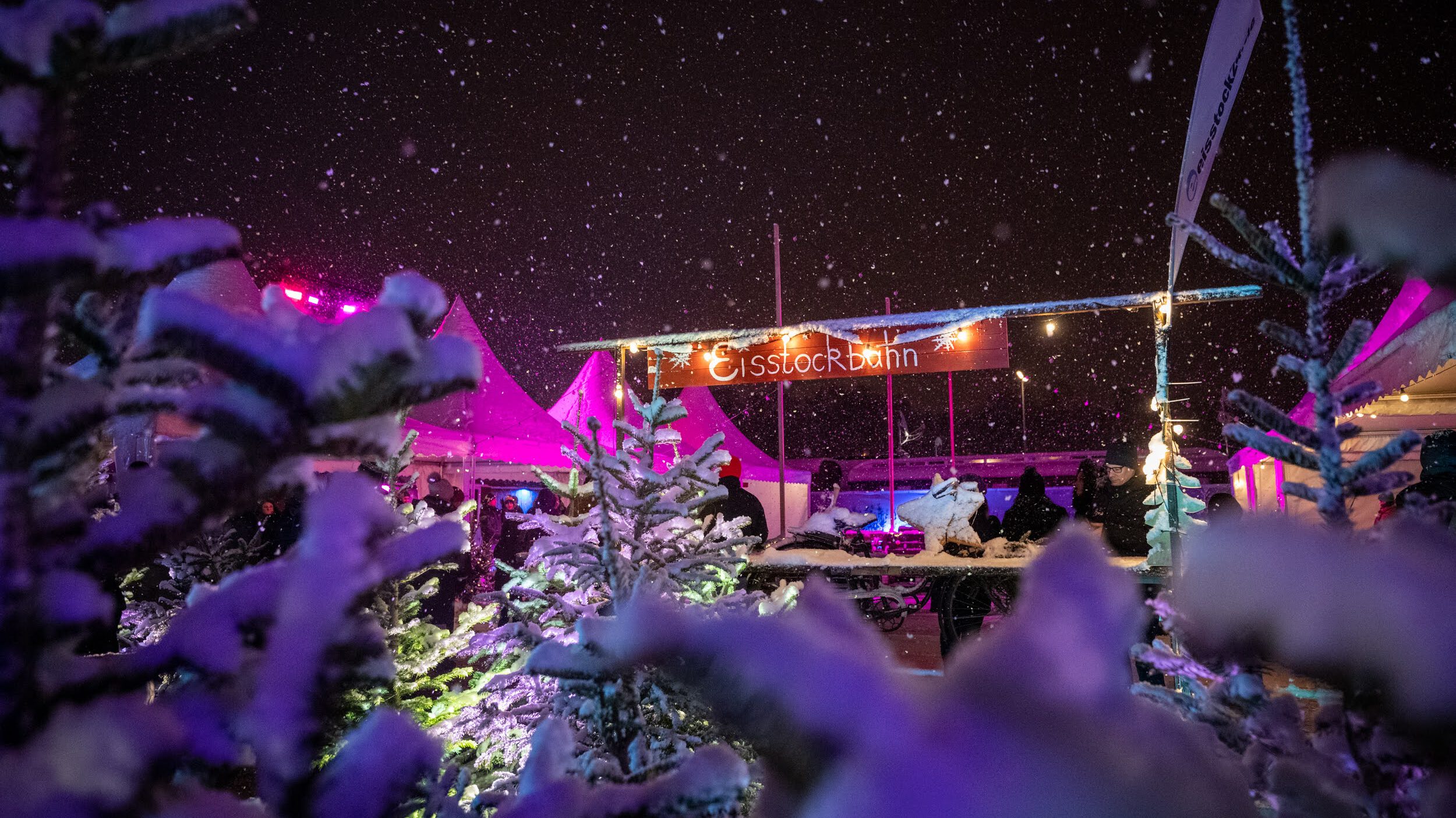 Eisstockschießen als Weihnachtsfeier ist Trend. Das Bild zeigt die Eisstockbahn auf dem Tollwood Winterfestival 2022 im Schneetreiben bei Nacht.
