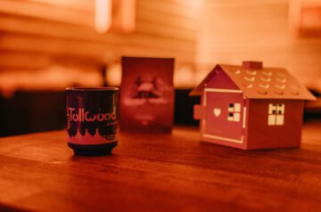 Das Bild ist von einer Weihnachtsfeier 2023 im Hexenkessel auf dem Tollwood Winterfestival und zeigt eine Glühweintasse, ein Hexenhäuschen und eine Menükarte auf einem Holztisch. Der Hintergrund ist warm ausgeleuchtet.