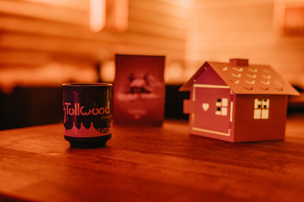 Das Bild ist von einer Weihnachtsfeier im Hexenkessel auf dem Tollwood Winterfestival und zeigt eine Glühweintasse, ein Hexenhäuschen und eine Menükarte auf einem Holztisch. Der Hintergrund ist warm ausgeleuchtet.
