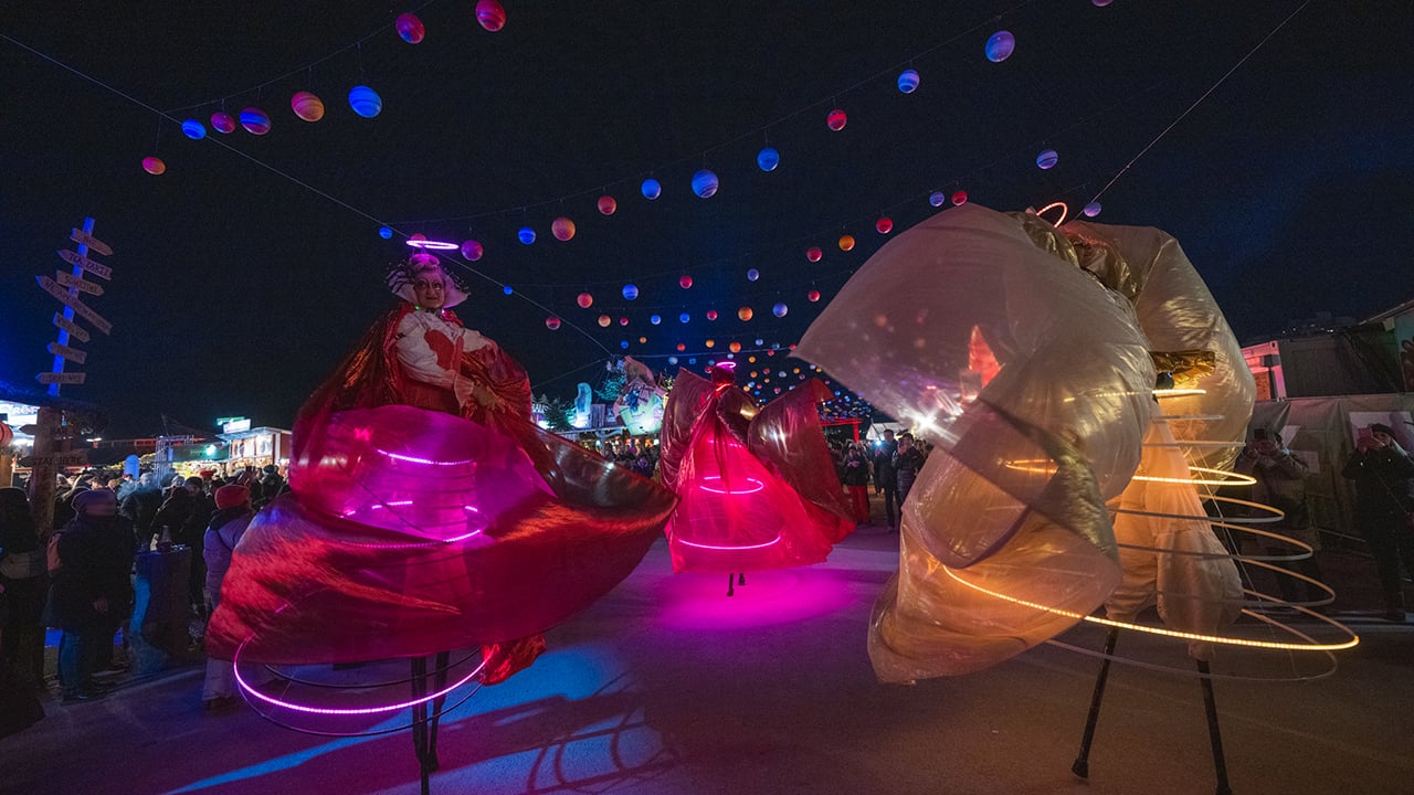Geheimtipp Weihnachtsfeier 2024: Das Bild zeigt die Performance Künstler*innen auf dem Tollwood Winterfestival vor dem Hexenkessel. Zu sehen sind drei Stelzenläufer*innen mit Kostümen aus weiten Tüchern und leuchtenden Ringen. Über den Köpfen der staunenden Besucher*innen und der Künstler*innen sieht man eine Planeteninstallation.