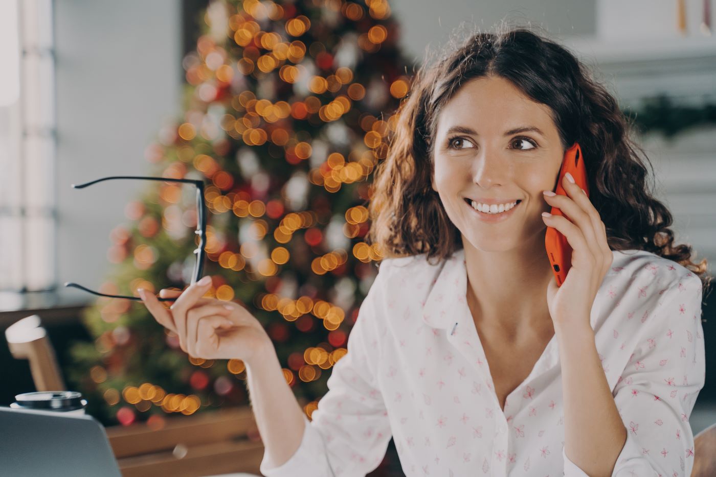 Das Bild zum Thema "Freibetrag Weihnachtsfeier 2024" zeigt eine jung hübsche Frau beim Organisieren. In der rechten Hand hält sie ihre Brille und mit der Linken ihr Mobiltelefon an ihr Ohr. Im Hintergrund ist ein Weihnachtsbaum zu erkennen. Ihr Lächeln lässt vermuten, dass sie ein erfolgreiches Gespräch führt.