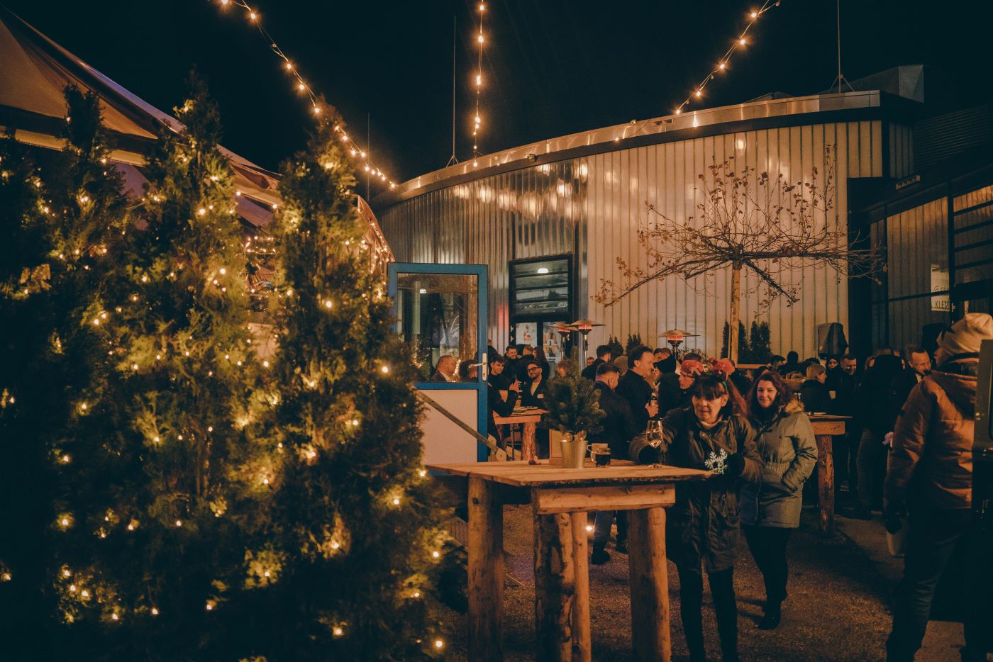 Eine Location für Ihre Weihnachtsfeier ist die Jochen Schweizer Arena allemal. Das Bild zeigt die Gäste einer Firmenweihnachtsfeier vor einer traditionellen Almhütte am rustikalen hölzernen Stehtischen. Im Hintergrund die moderne Eventanlage mit dem Flugkanal.