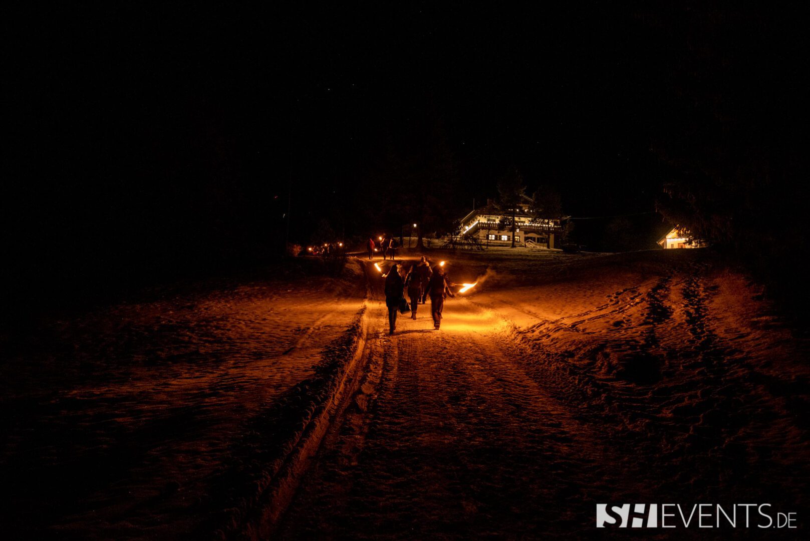 Eine Gruppe von Menschen wandert in der Dunkelheit mit Fackeln durch eine verschneite Winterlandschaft. Der Weg ist in warmes Licht getaucht und führt zu einer hell erleuchteten Berghütte im Hintergrund. Die Szene vermittelt winterliche Gemütlichkeit und festliche Stimmung – ideal für ein HüttenEvent als Weihnachtsfeier.