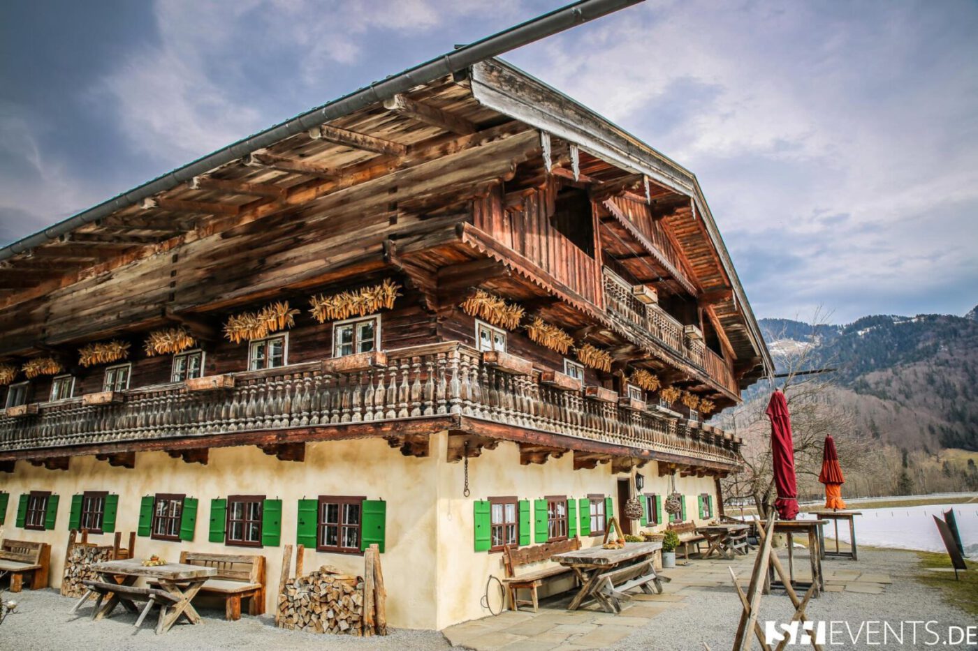 Traditionelles Bauernhaus in den bayerischen Bergen mit Holzbalkon, grünen Fensterläden und rustikaler Außengastronomie.